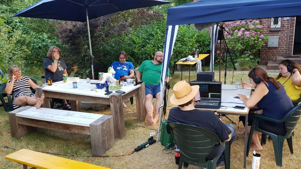 Auf dem Bild sind sieben Menschen zu sehen. Drei von ihnen sitzen rechts unter einem Pavillion, Gesche und Abby Podcasten dort, Jörn unterstützt mit der Tontechnik. Die anderen Gäste sitzen an einem Tisch und hören zu. Außer Rüdiger, der hat das Foto geschossen.